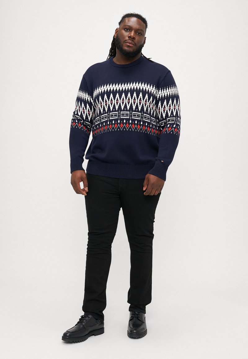 Navy knitted sweater with a white, red, and black geometric pattern, ribbed cuffs and hem, paired with black trousers and black shoes.