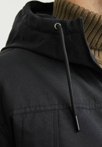 Chaqueta negra con capucha, con un cuello de punto marrón texturizado, que presenta una cremallera, cordón ajustable y un acabado de tela suave.