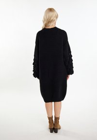 Černé pletené oversized svetr-šaty s texturovanými rukávy a zakulaceným lemem, zkombinované s hnědými kotníkovými botami na blokovém podpatku.