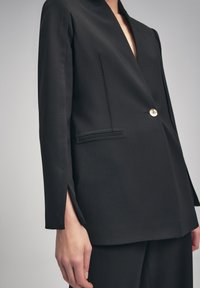 Blazer negro con solapas muestradas, cierre de botones dorados y aberturas laterales en las mangas. Corte ajustado con líneas limpias y una textura suave.