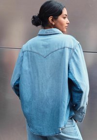 Giacca di denim di colore azzurro chiaro, vestibilità rilassata, con un design a yoke appuntito, chiusura con bottoni e orlo leggermente sfilacciato lungo le maniche e il fondo.