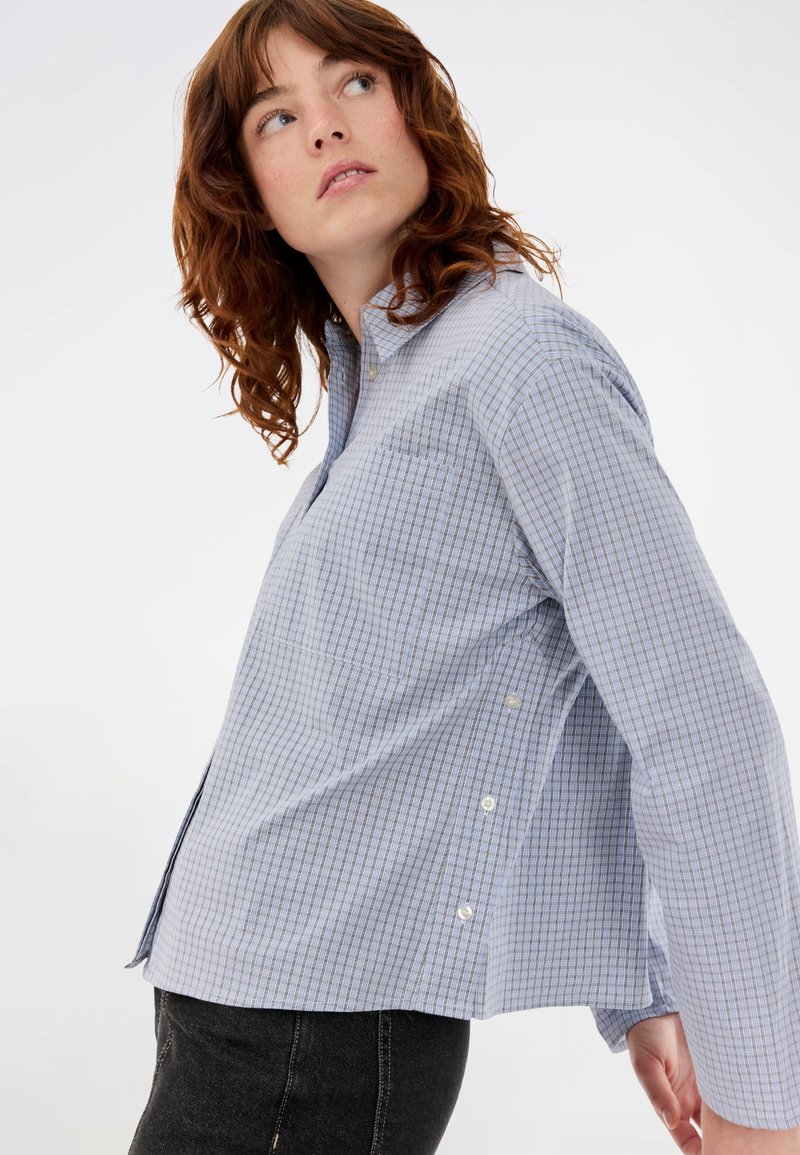 Νεαρή γυναίκα με σγουρά κόκκινα μαλλιά, φορώντας ένα μπλε καρό πουκάμισο και μαύρα παντελόνια, κοιτάζει προς τα αριστερά ψηλά.