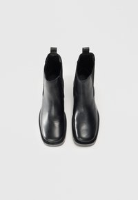 Botas de cuero negras hasta el tobillo con una textura suave, punta cuadrada, paneles laterales elásticos y tiradores para facilitar el uso. La suela es plana y resistente.