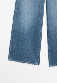 Niebieskie jeansy z denimu o szerokich nogawkach i widocznym przeszyciu na białym tle.