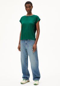 T-shirt vert à manches courtes en tissu lisse, associé à un jean bleu clair ample et des baskets blanches avec des accents verts.