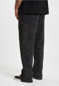 Store svarte baggy denimbukser med en falmet tekstur og preget hodeskalle-logo. Har sidelommer med glidelås og en løs passform. Rent, minimalistisk design.