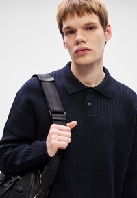 Pólo naval com nervuras, com um decote com colarinho e uma placket de três botões. O modelo segura uma mala preta de ombro com uma alça.