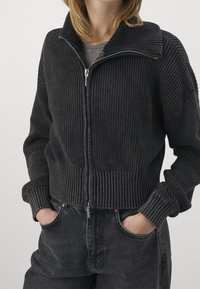 Schwarze gerippte Strickjacke mit Reißverschluss, breitem Kragen und langen Ärmeln, aus strukturiertem Stoff und mit tailliertem Bündchen. Dazu schwarze Jeans.