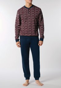 Uomo che indossa una maglietta a maniche lunghe color marrone bordeaux con motivo e pantaloni blu navy liscio, in piedi a piedi nudi contro uno sfondo bianco semplice.