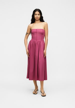 Vrouw met lang donker haar staat in een strapless magenta midi-jurk en zwarte flipflops, tegen een egale witte achtergrond.