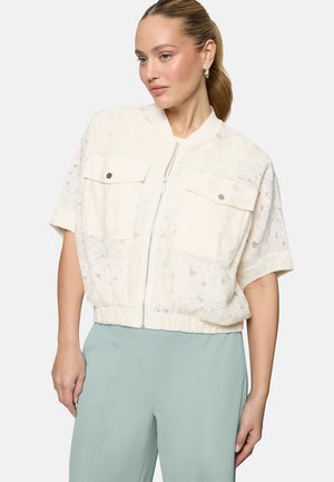 Femme portant une veste blanche à manches courtes en dentelle avec fermeture éclair, poches avant et pantalon bleu clair, posant avec la main reposant sur la cuisse.