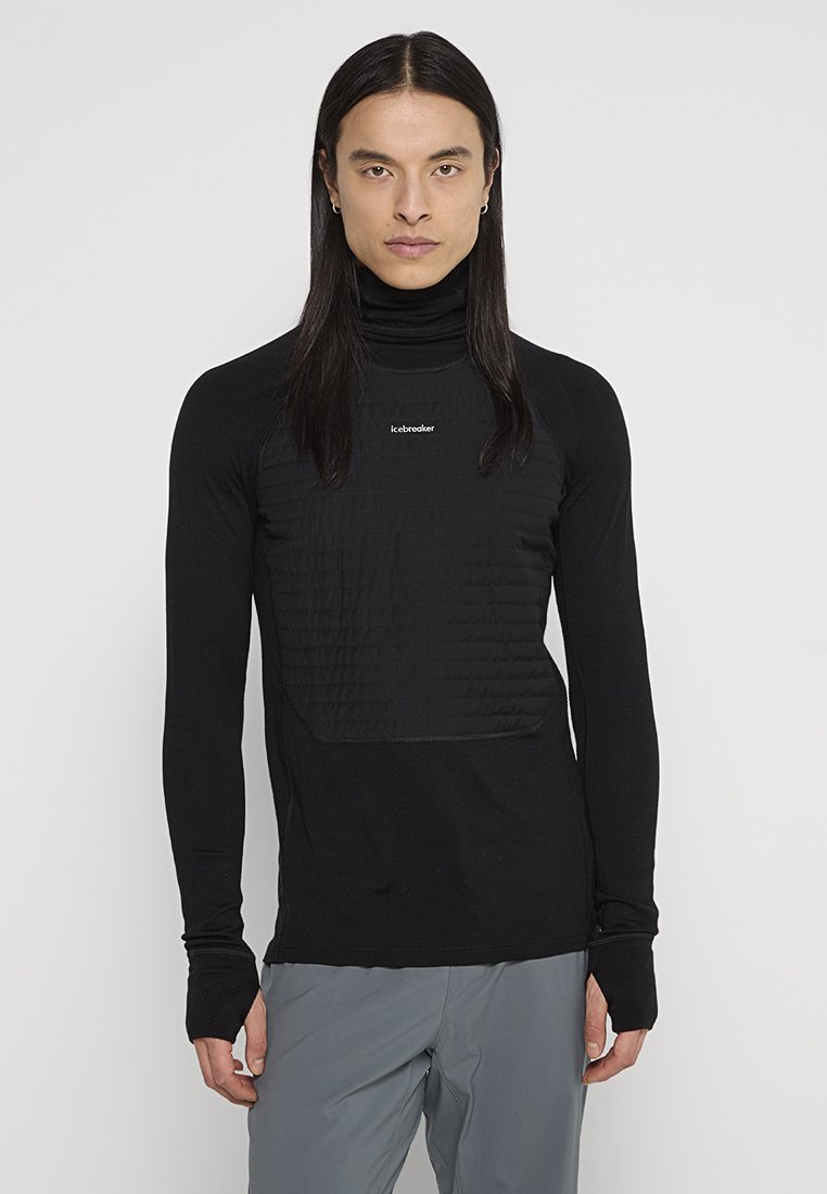 Erwachsene Person mit langem, dunklem Haar, die einen schwarzen, gesteppten Rollkragenpullover und graue Hosen trägt und vor einem schlichten weißen Hintergrund steht.