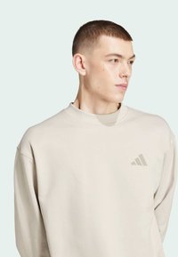 Sudadera beige con cuello redondo, que presenta un acabado texturizado y un sutil logo de Adidas en el lado izquierdo del pecho. Corte holgado y puños acanalados.