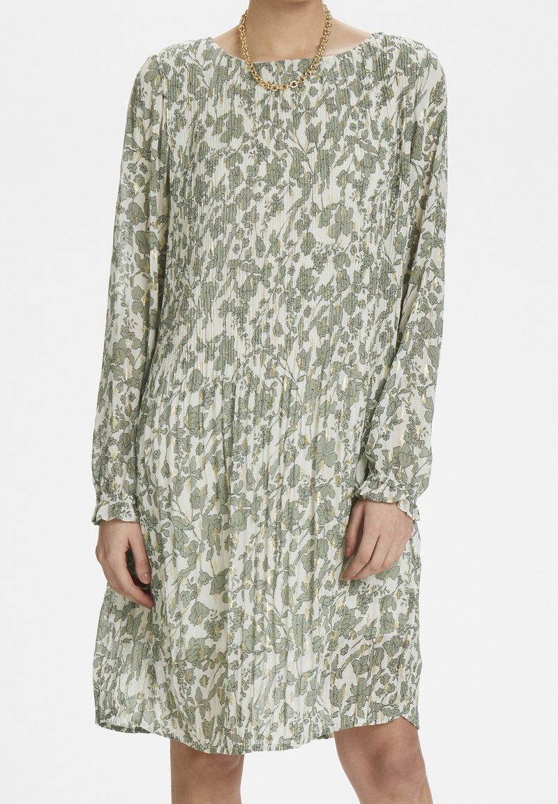Femme portant une robe plissée à manches longues, longueur genoux, avec un motif floral vert sur un tissu blanc et un collier en chaîne doré.