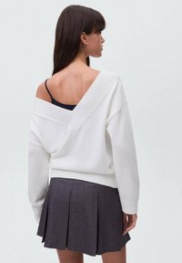 Bela pletena pulover z odkrito ramo in ribastimi poudarki, združena s plisirano temno sivo krilo. Pulover ima globok V-izrez na hrbtu.