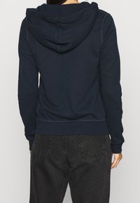Felpa blu navy con un ampio cappuccio, caratterizzata da una texture liscia e polsini a costine. Sono visibili dettagli di cucitura lungo le cuciture.