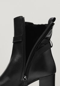 Zwarte leren enkelboot met een gedeeltelijk open zijrits, die de zachte binnenvoering en de gestapelde blokhak toont.