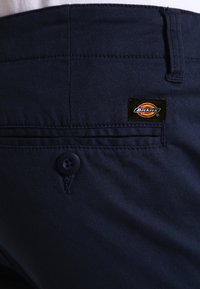 Pantalons en coton bleu foncé avec une poche arrière portant un logo Dickies noir et rouge, une fermeture à bouton et des détails cousus.