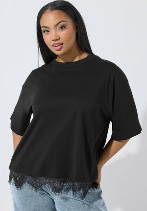 Ung kvinde med langt sort hår iført en løs sort top med blondekant og lyseblå jeans mod en lys baggrund.