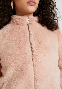 Jas van imitatiebont in zachtroze, met een hoge kraag, een volledige ritssluiting en een zachte, gestructureerde oppervlakte. Zilverkleurige accenten benadrukken het ontwerp.