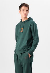 Felpa verde in cotone con tasca frontale, cappuccio regolabile e logo arancione. Abbinata a pantaloni da jogging verdi con toppa del logo.