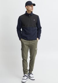 Pullover in pile blu navy e nero con mezzo zip, abbinato a pantaloni cargo oliva dotati di molteplici tasche e sneaker bianche con dettagli neri.