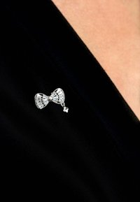 Silver brooch shaped like a bow, featuring intricate geometric patterns and a dangling charm, pinned to black fabric.