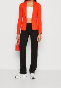 Blazer rosso aderente, top bianco corto, pantaloni neri e sneakers bianche. Tiene una piccola borsa rossa. Focalizzazione su colori vivaci e design moderno.