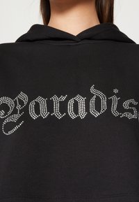 Sweat à capuche noir avec un motif en strass argenté formant le mot "Paradise" en police ornée sur le devant. Tissu doux avec une coupe décontractée.