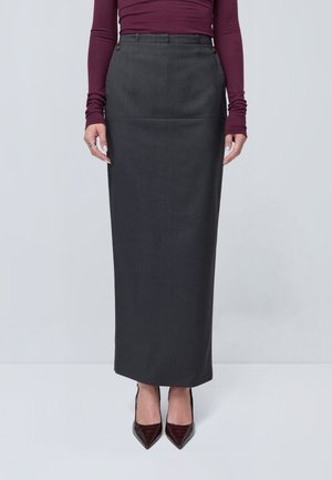 Femme portant une jupe crayon longue gris foncé, un haut bordeaux à manches longues et des talons pointus foncés, debout devant un fond clair uni.