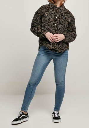 Person, die ein leopardgemustertes Hemd mit Knöpfen, blaue Skinny-Jeans und schwarz-weiße Sneakers trägt, steht mit verschränkten Händen.