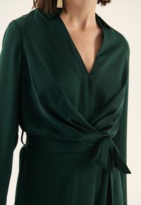 Robe portefeuille verte avec encolure en V, manches longues et ceinture nouée à la taille. Fabriquée en tissu lisse et léger avec un léger éclat.