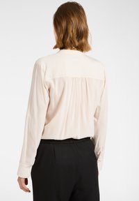 Jasno-beżowa bluzka z długimi rękawami i plisowanym tyłem. Materiał wydaje się gładki z lekko połyskującą fakturą, dopełniony czarnymi spodniami.