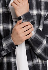 Camicia di flanella a scacchi bianco e nero con polsini abbottonati, caratterizzata da una texture morbida e due tasche frontali. Sotto, una t-shirt bianca.