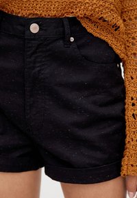 Shorts in denim nero con macchie colorate, dotati di chiusura con bottone e orlo arrotolato. Il tessuto è liscio con una leggera lucentezza.
