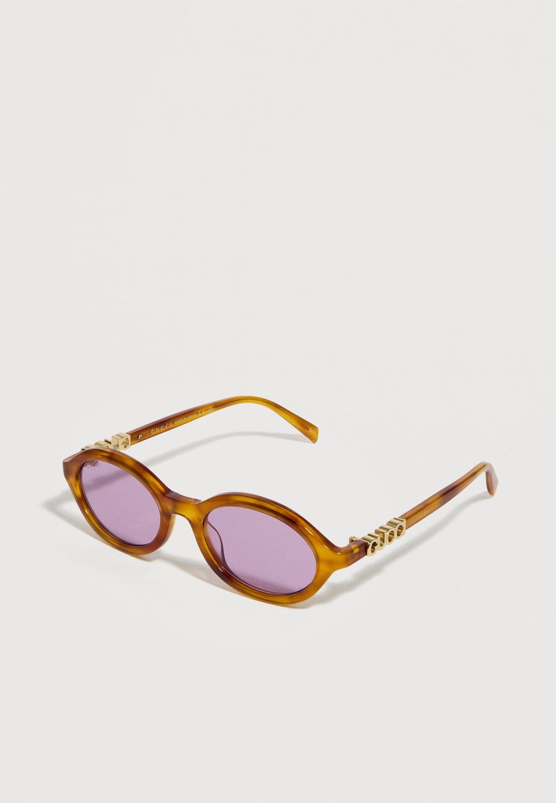 Lunettes de soleil ovales en écaille de tortue marron avec des verres violets et des accents décoratifs dorés sur les branches, reposant sur une surface blanche.