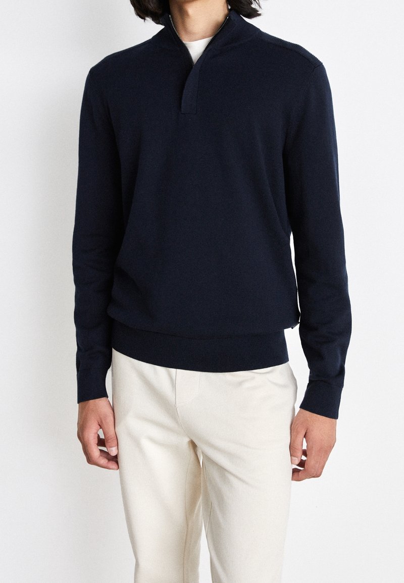 Pull-over en maille bleu marine avec col à fermeture à glissière partielle, poignets et ourlet côtelés, associ é à un pantalon beige clair, créant un look élégamment décontracté.