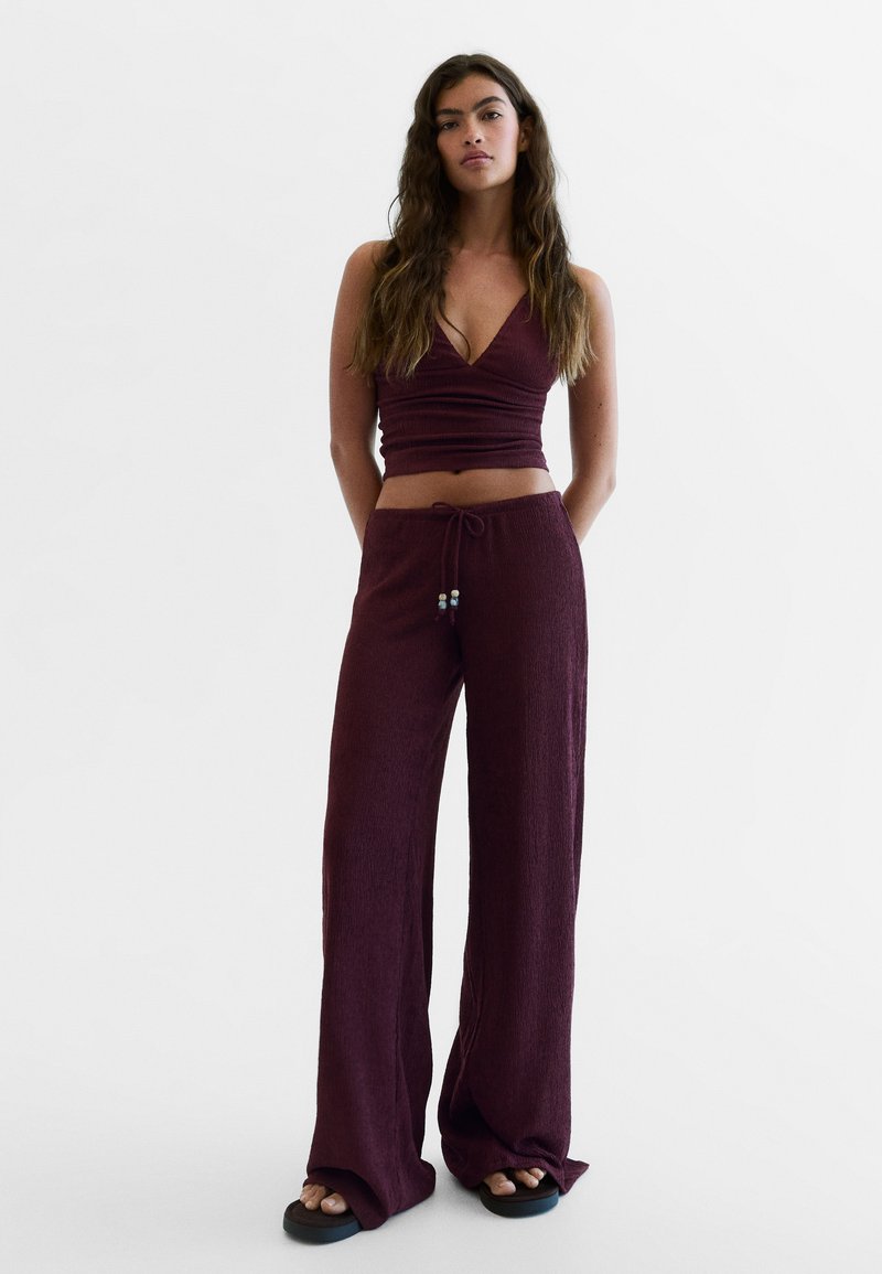 Femme aux longs cheveux portant un crop top bordeaux sans manches à col en V profond et un pantalon large assorti, debout, les mains dans le dos.
