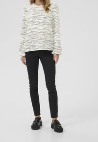 Biała bluzka z długim rękawem i czarnym falistym wzorem, zebranymi mankietami, zestawiona z czarnymi skinny jeansami i czarnymi platformowymi loafersami. Prosty, minimalistyczny design.