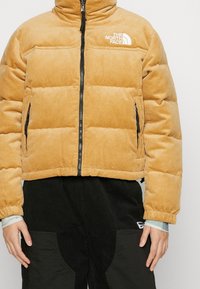 Person som bär en beige kordflöjel North Face puffjacka med svart dragkedja och svarta byxor med fickor synliga på sidorna.