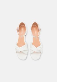 White sandals with a knotted fabric design, open toe, and ankle strap with a buckle. Smooth texture and light tan insole.