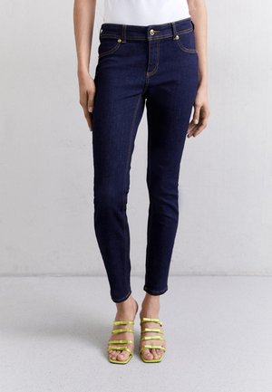 Donna che indossa jeans skinny blu scuro e sandali con tacco alto verdi brillanti con cinturini, in piedi su un pavimento grigio chiaro.