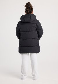 Mujer vistiendo un abrigo acolchado con capucha negra y pantalones de chándal gris claro, de pie de espaldas sobre un fondo blanco.