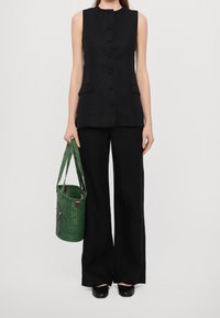 Černý top bez rukávů s knoflíky vpředu a bočními kapsami, doplněný volnými černými kalhotami a zelenou pletenou taškou na rameno s motivem palmy.