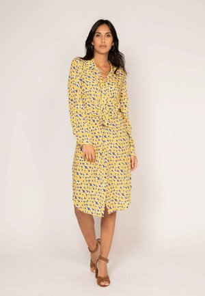 Robe à manches longues, boutonnée, de couleur jaune vif avec un motif abstrait bleu, dotée d'une taille à nouer et d'une fente à l'avant. Le tissu semble léger.