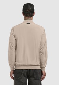 Beige letvægts bomberjakke med ribstrikkede manchetter og kant, glat stof, og et lille sort mærke ved halsen, set fra bagsiden.