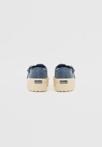 Denim anyagú, slip-on cipők bézs gumitalppal, hátul pánttal és logó részletekkel a sarkán. Hétköznapi, lapos profilú dizájn.