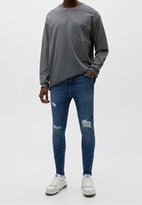 Šedá košile s dlouhým rukávem a náprsní kapsou, spárovaná s těsnými modrými džíny s opotřebovanými detaily a bílými teniskami.