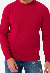 Pull rouge à col rond avec manches longues, poignets et ourlet côtelés. Associé à un jean bleu déchiré. Texture lisse et coupe ajustée.