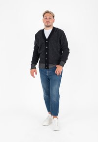 Cardigan a maglia grigio scuro con scollo a V, chiusura con bottoni neri e polsini grigi a contrasto. Indossato su una t-shirt bianca e abbinato a jeans blu.